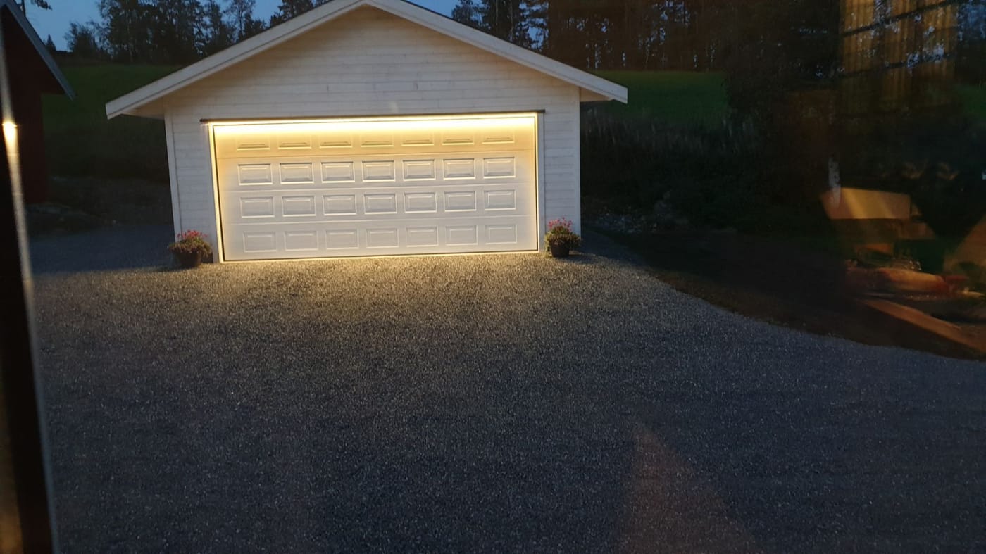 Hvit garasje med LED-lys rundt porten i skumringen, som lyser opp grusen foran.