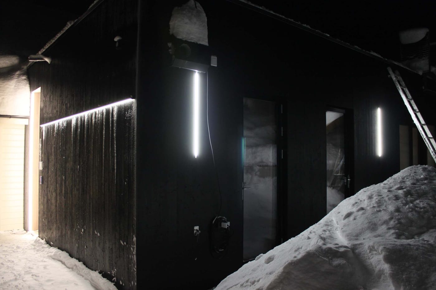 Mørk trefasade med LED-lys langs veggen og ved dørene en vinterkveld, med snø og istapper.